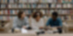 Three people sit on a couch in a library, discussing books on a table. Shelves filled with books are in the background. Mood is focused.